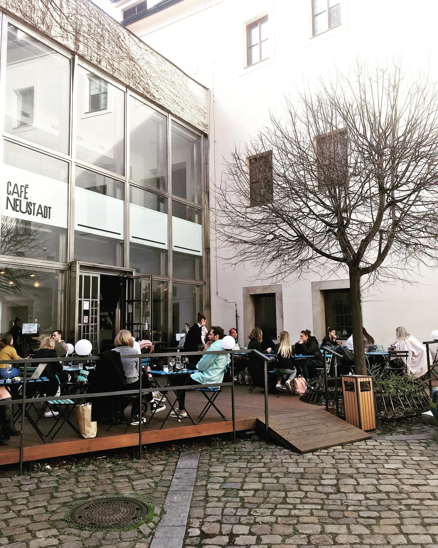 Terasa Café Neustadt na dlážděném nádvoří Novoměstské radnice, modré bistro židle před prosklenou fasádou s nápisem CAFÉ NEUSTADT